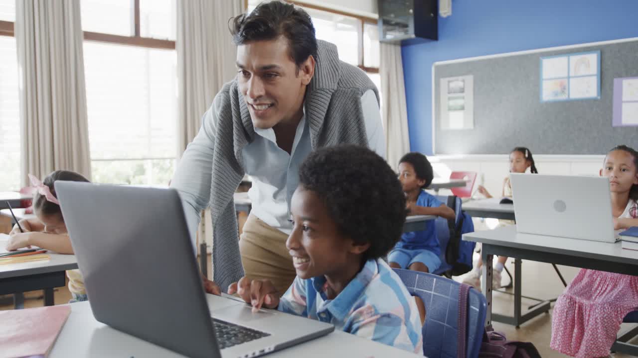 Happy diverse male teacher helping boy at desk using laptop in elementary school class, slow motion