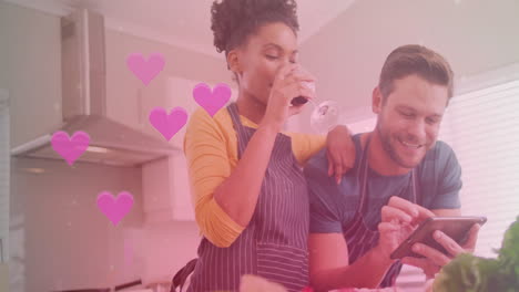 Animation-of-hearts-over-diverse-couple-cooking-together-and-using-tablet-in-kitchen-at-home