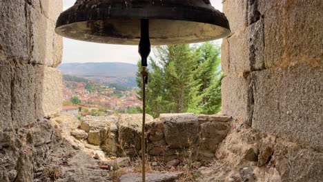 filming inside a bell tower where we see a large stained bronze bell, the clapper and its rope appear, we see the stone walls and the floor with earth and plants, and in the background a town appears