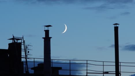 Time-lapse-of-beautiful-waxing-crescent-moonset-with-fast-moving-clouds-over-the-roof-with-chimneys,-medium-shot