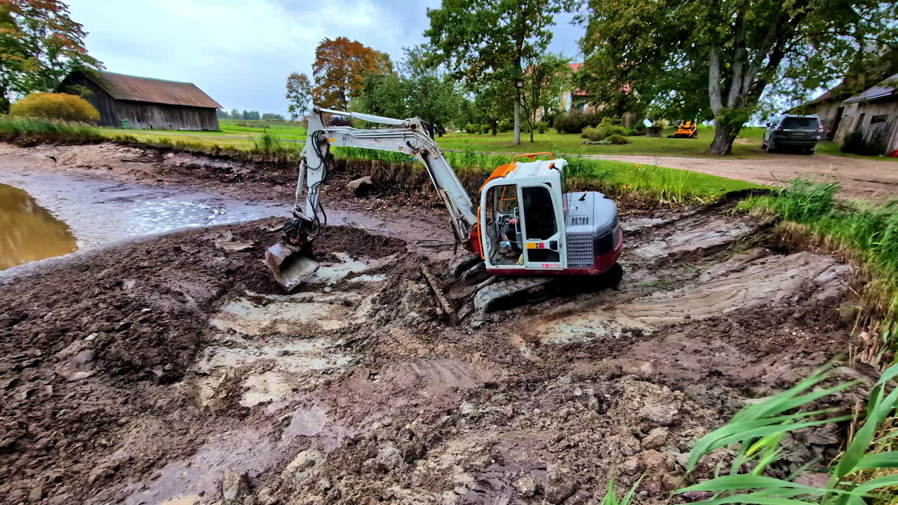 Mechanical Excavator Digging Pond Near Rural Village. Panning Shot