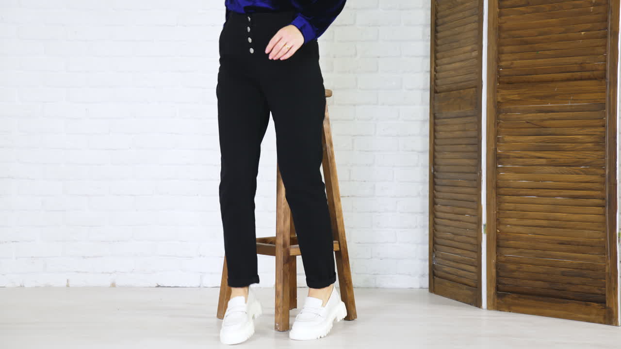 Woman in black trousers demonstrates white shoes. Lady comes and sit on the high wooden stool. White wall backdrop.