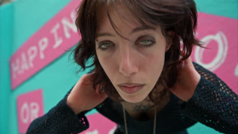 outdoor fashion portrait of young alternative style woman leaning in towards camera checking hair against graffiti covered walls of london city street uk in real time