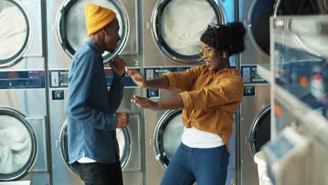 young cheerful pretty girl and stylish handsome guy having fun and dancing in laundry service room while machines washing on background