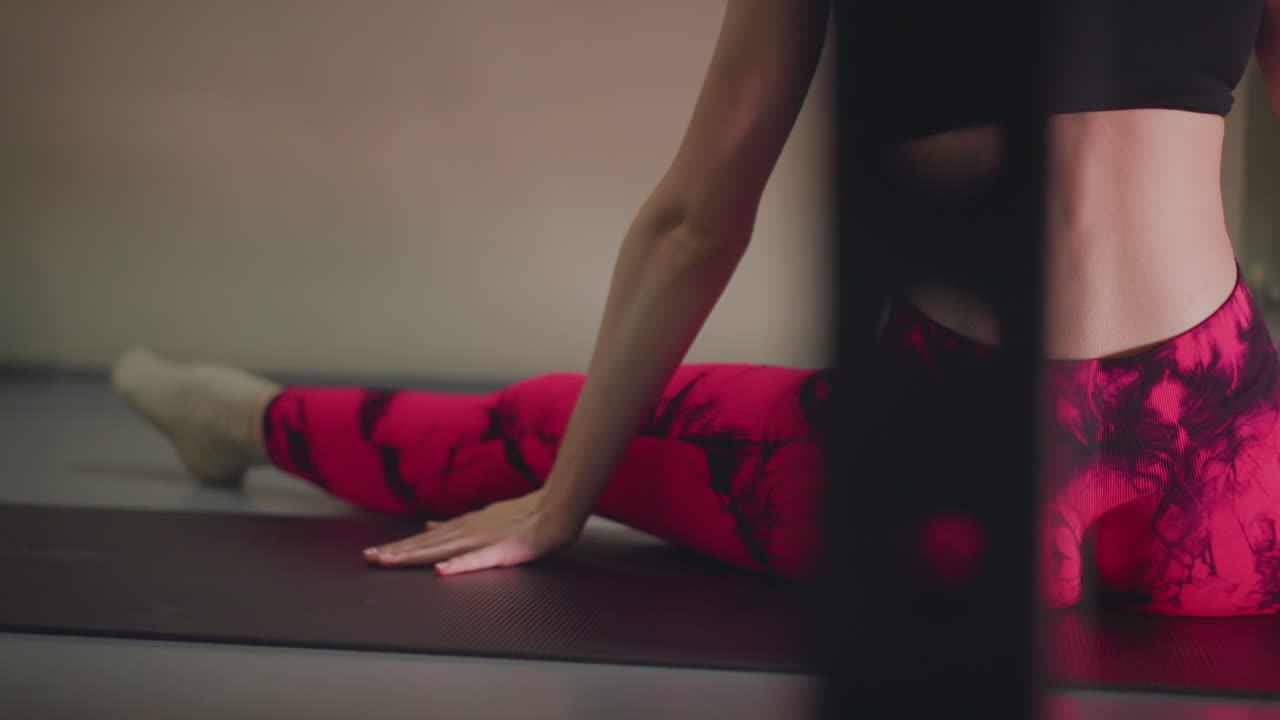 Donna su tappetino che esegue spaccate da seduta, leggings rosa e crop top nero, focus su zona lombare e glutei, illuminazione da studio interna, lenta e intenzionale apertura delle anche e attivazione dei glutei per flessibilità e postura.