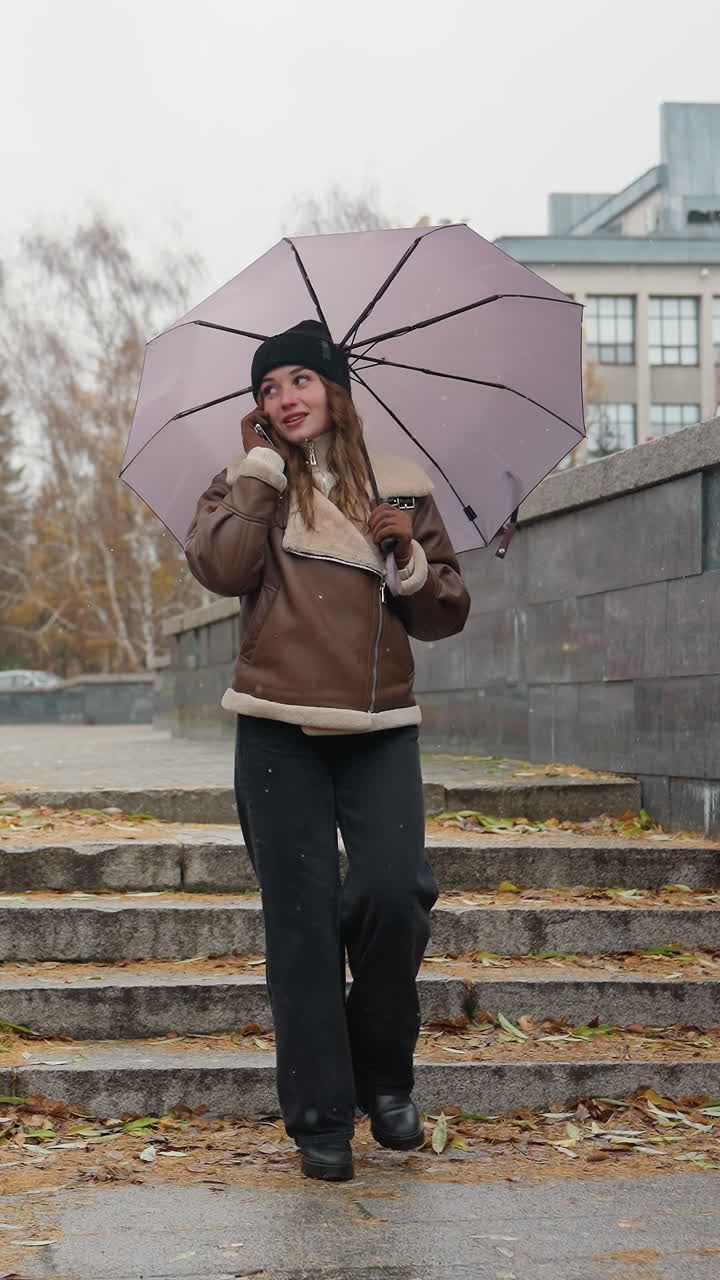 Ragazza sorridente che fa una telefonata mentre tiene un ombrello, indossa un berretto di lana nero, una giacca shearling marrone e pantaloni neri, cammina in un parco urbano in una fredda giornata nuvolosa con una leggera nevicata