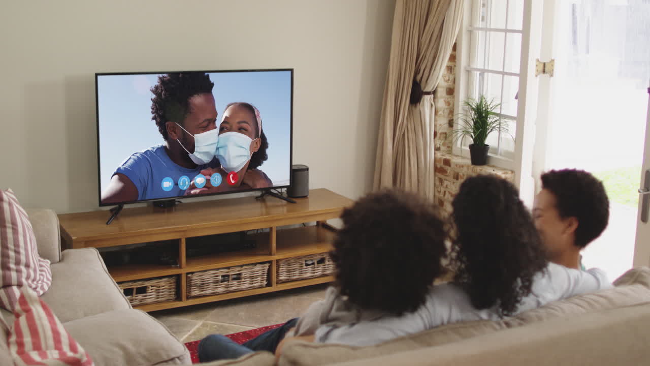Famiglia afroamericana vista da dietro in videochiamata sulla TV, seduta sul divano a casa.