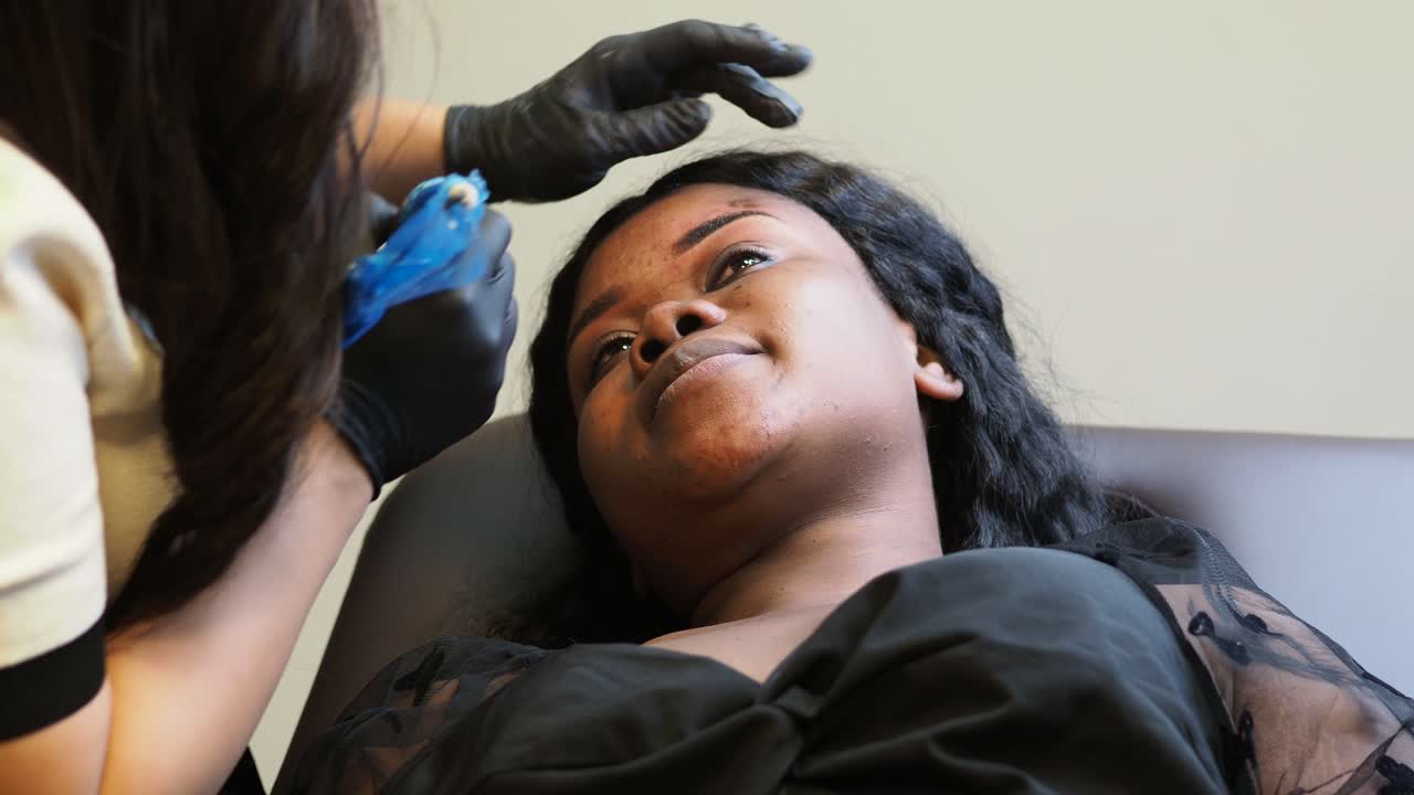 Beautician wearing black gloves is using a rotary tattoo machine for applying permanent makeup on a customer's eyebrows