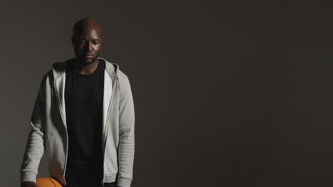 Studio-Portrait-Shot-Of-Male-Basketball-Player-Wearing-Tracksuit-Holding-Ball-Against-Dark-Background