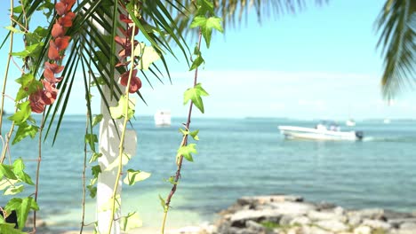 Schwenkaufnahme-Von-Blumen-Auf-Einem-Bogen-Mit-Einem-Vorbeifahrenden-Boot-Im-Hintergrund.-Hochzeitsort-Am-Strand-An-Einem-Sonnigen-Tag-Mit-Ruhe-Und-Frieden.-Sitzgelegenheiten-Im-Freien-Am-Sandstrand.-Verlobungssitzplätze-Mit-Tischen.