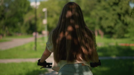 back view of young woman riding bicycle confidently through scenic park, long hair cascading down her back, shadows from trees creating patterns on her hair and outfit