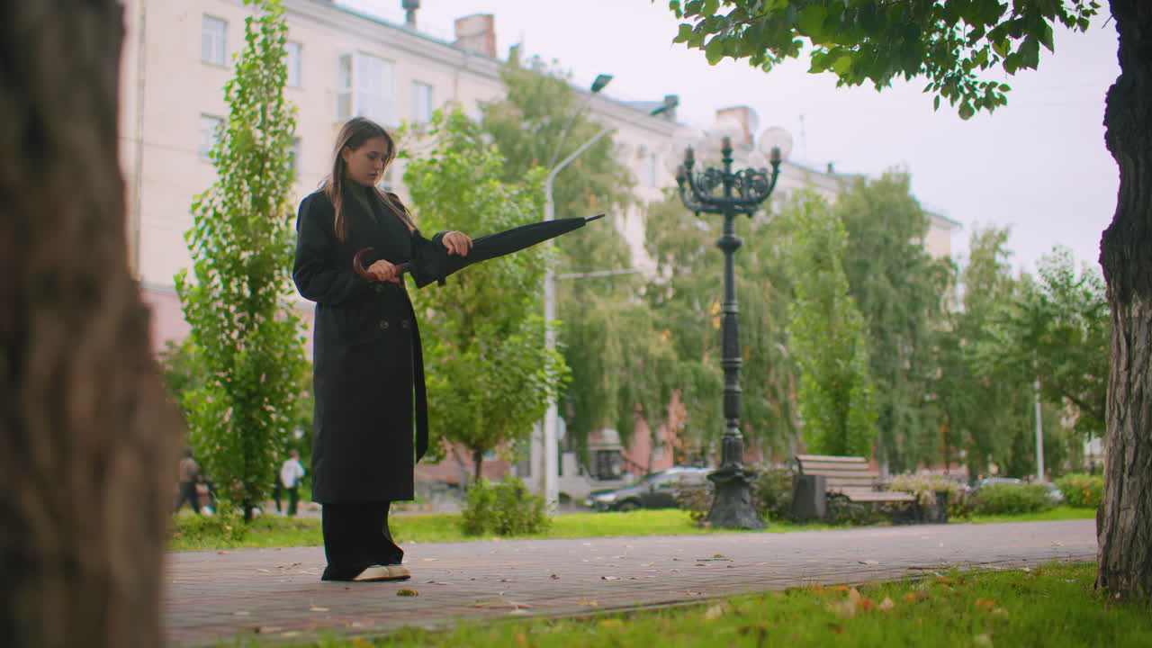Woman in long black trench coat standing on city park sidewalk holding closed umbrella, extending hand to check for raindrops, surrounded by trees, benches, lamp post, overcast autumn weather