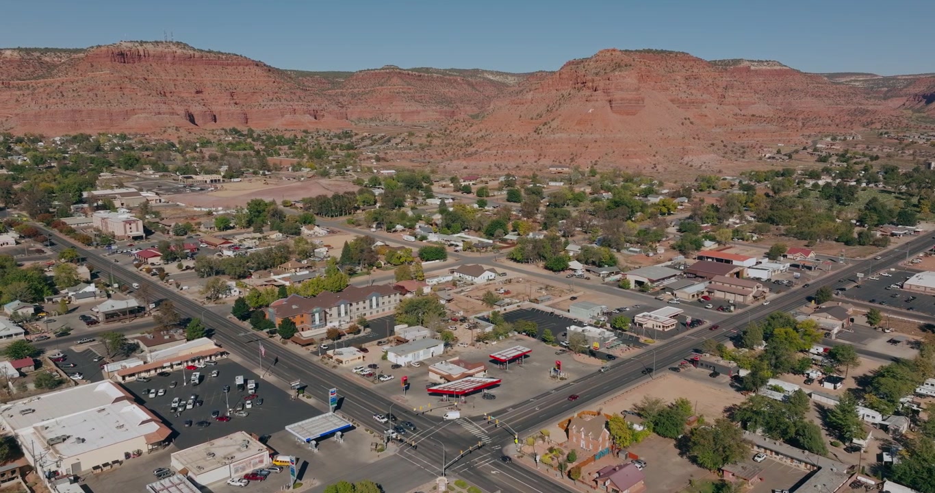 Quarteirões residenciais e ruas da cidade em Kanab, Utah, sob mesas de arenito vermelho, com drone em plano médio, acompanhando para a esquerda.