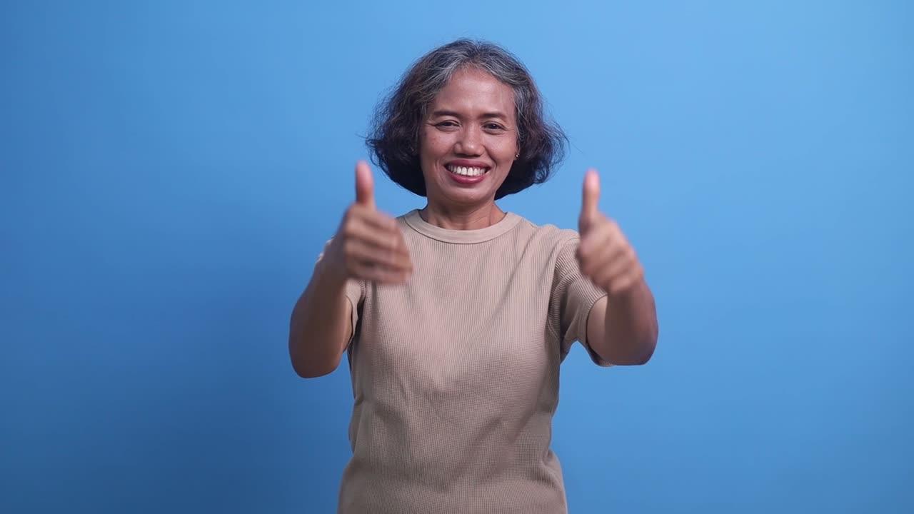 Satisfied enthusiastic Asian elderly woman accepting offer with thumbs up isolated on blue background