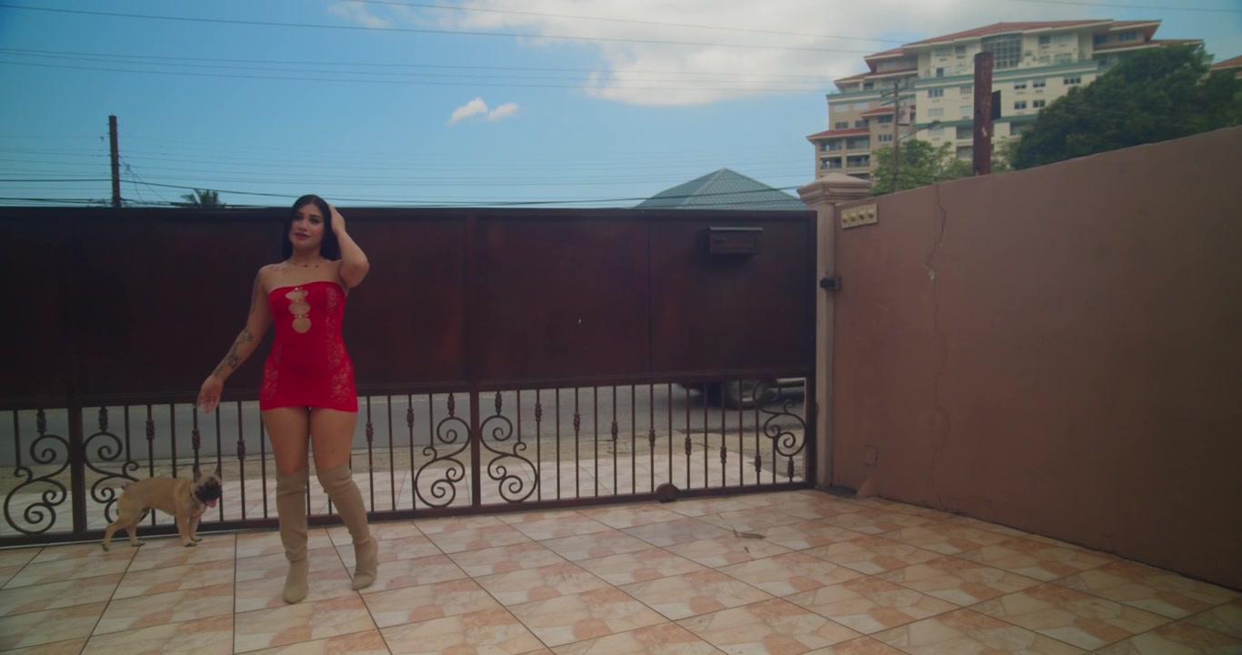 Movimento di telecamera frontale in tracking che mostra una donna latina sorridente che si rilassa su un divano modulare basso in lingerie rosso brillante, godendosi un momento giocoso e spontaneo