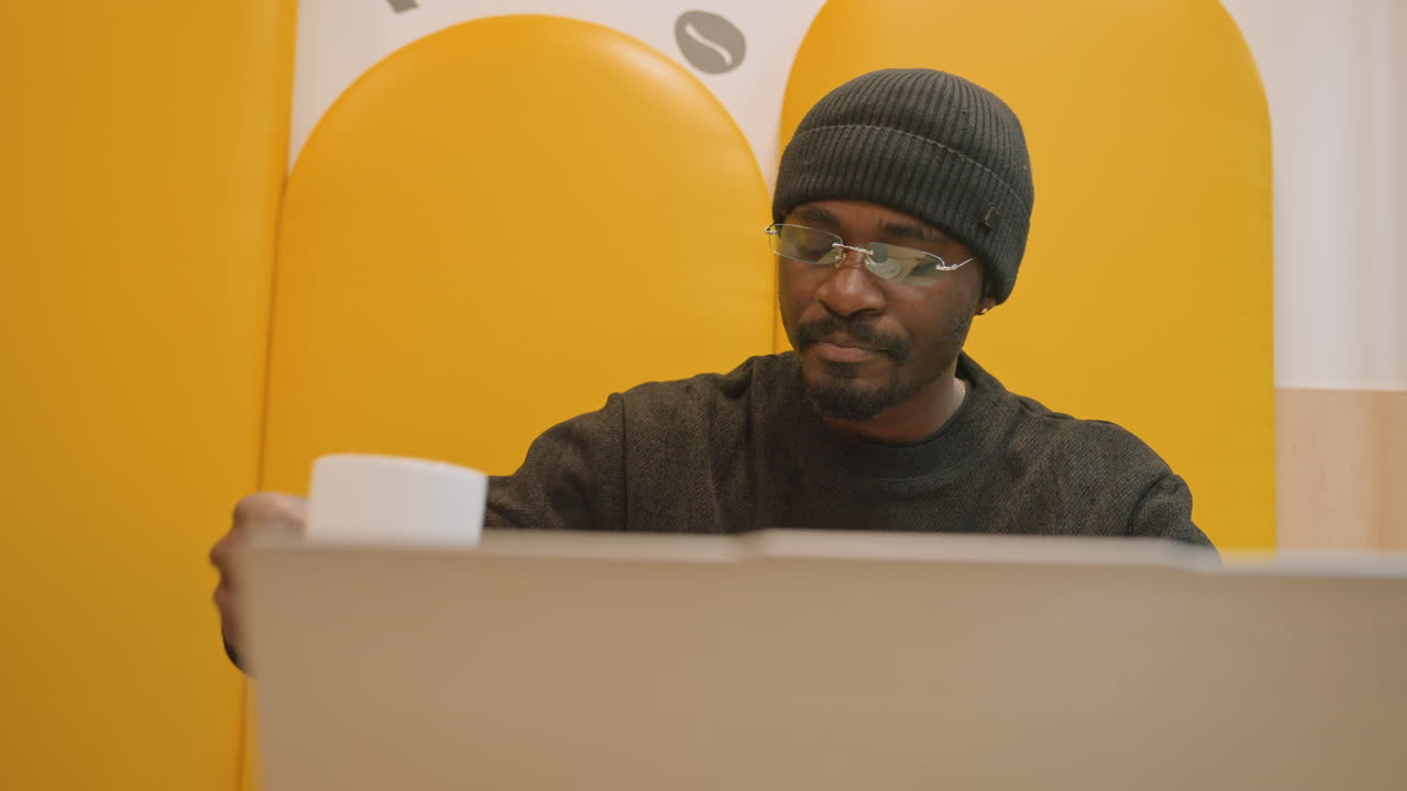 Man concluding work at laptop smiling, content creator reviewing finished project and sipping coffee, relaxed posture against warm yellow background, beanie and glasses, sense of achievement and calm