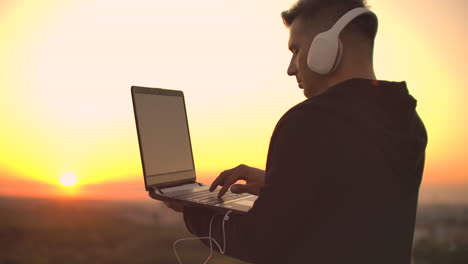 a man in headphones on the roof relaxes working remotely enjoying life despite a handsome kind of sipping beer and types on the keyboard. trade on the stock exchange using a laptop and enjoying the beautiful view