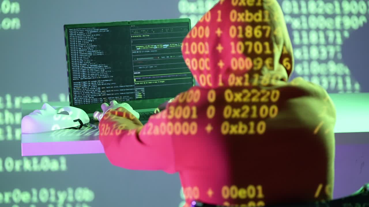A masked hacker in a red hoodie works on a laptop while yellow hex code illuminates the scene. A symbolic white mask rests beside, emphasizing themes of identity, privacy, and cyber manipulation