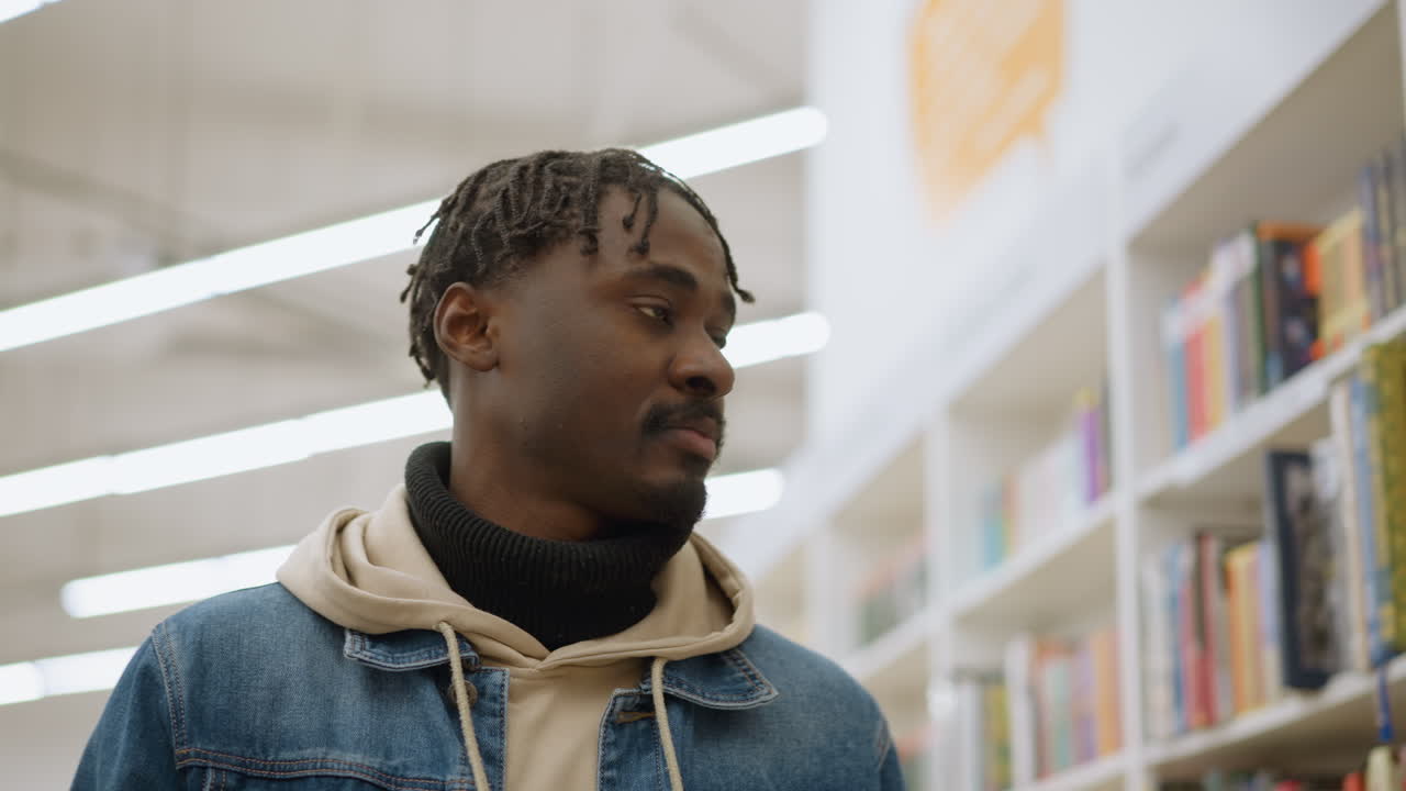 Man walking through bookstore aisle, wearing blue denim jacket and beige hoodie, browsing bookshelves with focus and interest, surrounded by colorful book spines in bright well-lit store