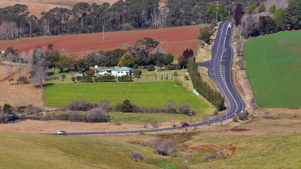 Drone ascends above winding rural highway, green fields, and farmhouses under soft natural daylight