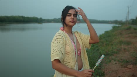 Woman-with-a-candy-cane-by-the-river-at-dusk,-wearing-a-sweater,-looking-back