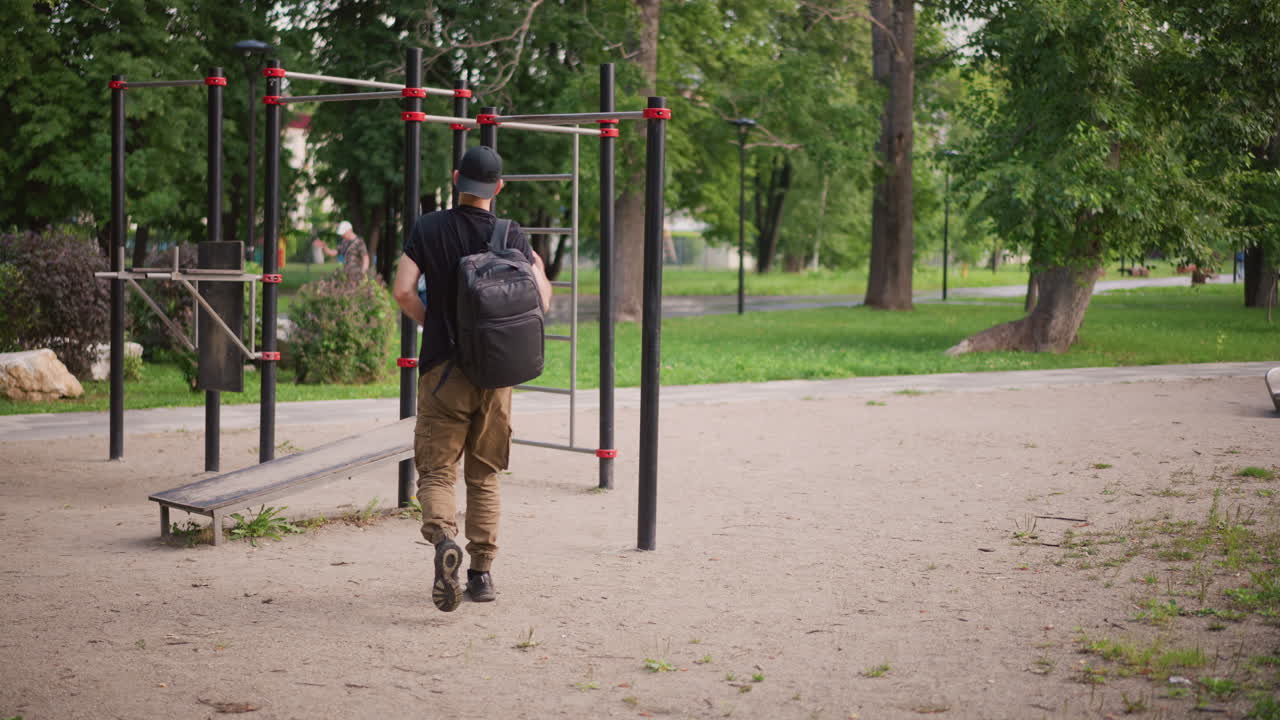 Individual Approaches Open Exercise Zone, Person Advances Into Outdoor Workout Space Confidently, An Athletic Man Carrying Gear Strides Purposefully Into Lush Outdoor Exercise Setting Early In Day