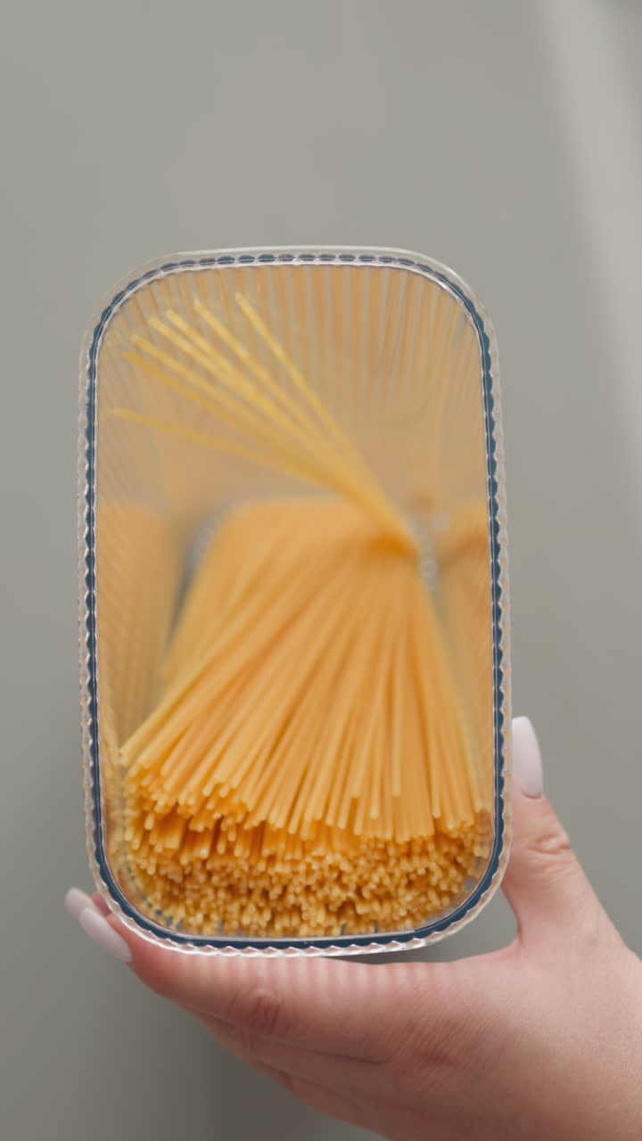 Woman puts spaghetti into transparent plastic container and closes lid shaking upper view. Young lady packs raw food into convenient box for storage