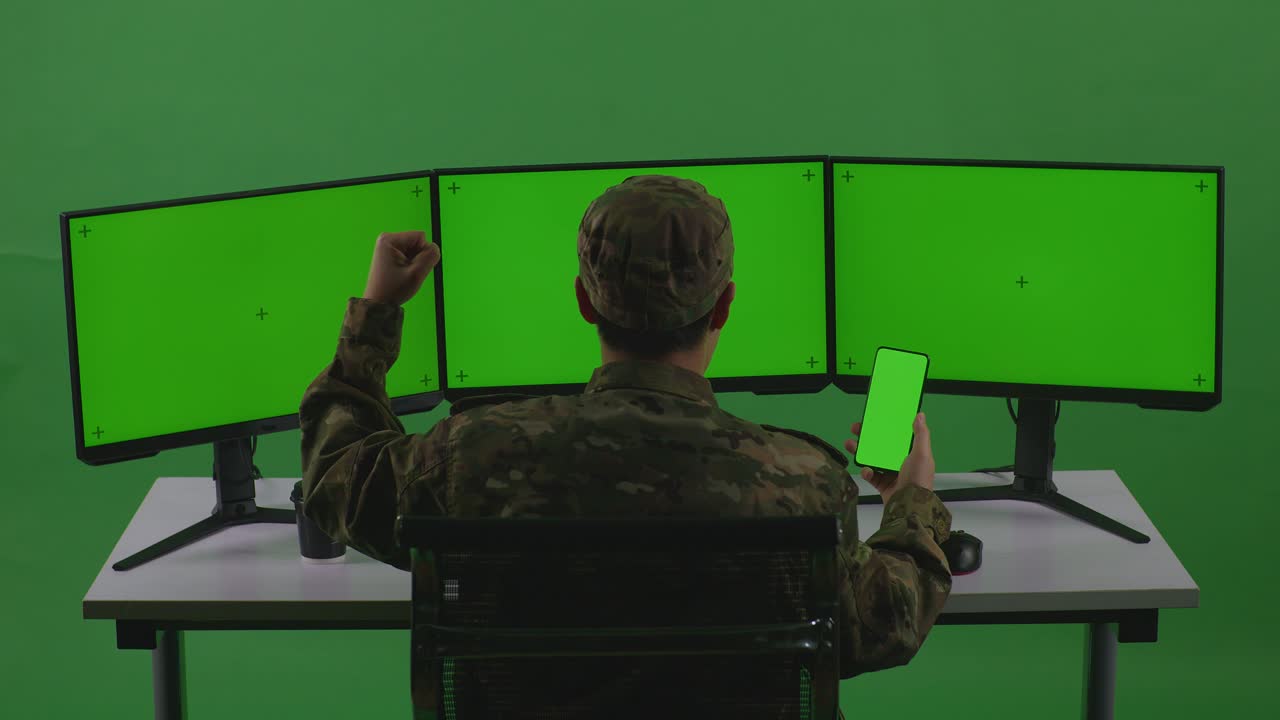 задний вид счастливого азиатского мужчины солдат смотрит на зеленый экран мобильного телефона и празднует во время работы с несколькими компьютерными мониторами в зеленом экране студии