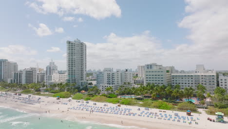 Modern-multistorey-apartment-buildings-or-hotels-on-sea-coast.-Backwards-reveal-of-sand-beach-with-loungers.-Miami,-USA