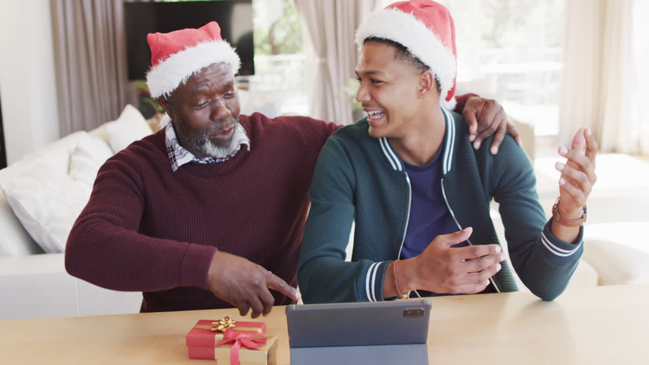 feliz padre afroamericano y hijo adulto con sombreros de navidad teniendo llamada de video de tableta, cámara lenta