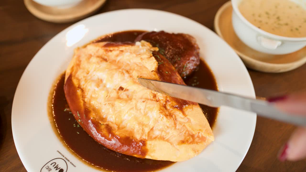 Hand slices open fluffy Japanese omelette rice with sauce in bright restaurant, overhead view