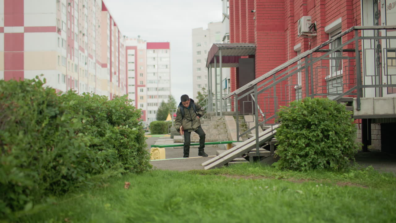 Rear view of kid carrying backpack and holding book walking toward bench, surrounded by greenery and residential buildings, leaves swaying gently in wind, urban lifestyle moment