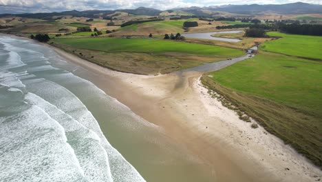 calm ocean waving on sandy beach, rolling hills of new zealand coastal farmland