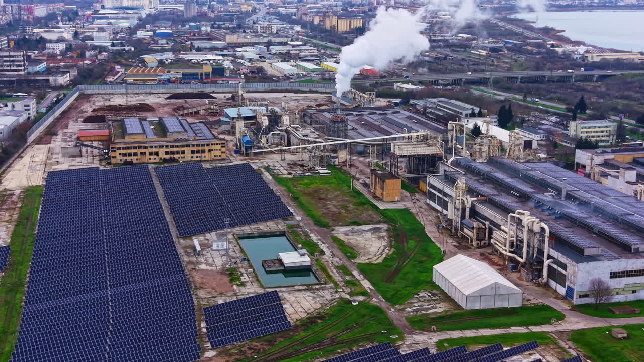 Power plant operates with solar panels nearby in an industrial area with factories and buildings in the background during daytime