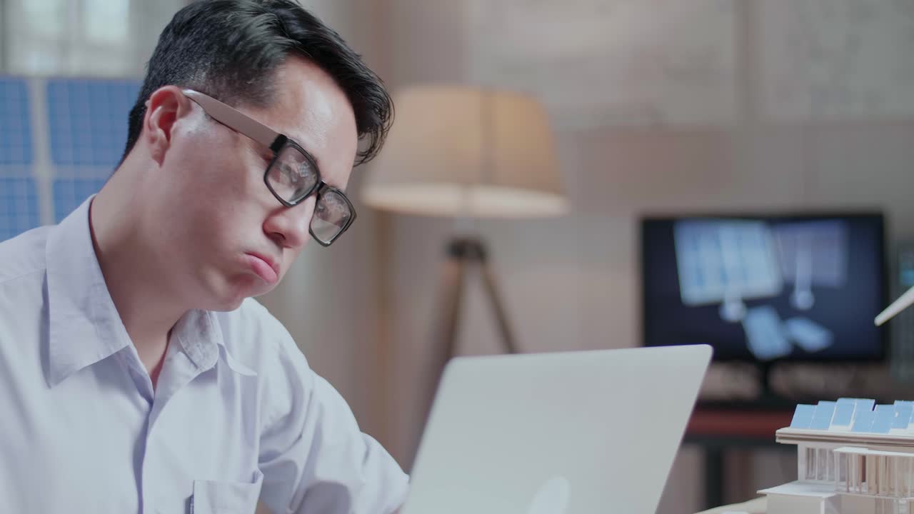 태양광 패널이 있는 작은 집의 모델과 함께 노트북 컴퓨터로 일하는 불만족한 아시아 남자의 클로즈업