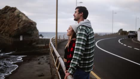 Woman-And-Man-Gazes-At-Ocean-Young-Couple-Shown-From-The-Side-Look-Out-Over-An-Ocean-In-Front-Of-The-Hills,-Talking,-Embracing