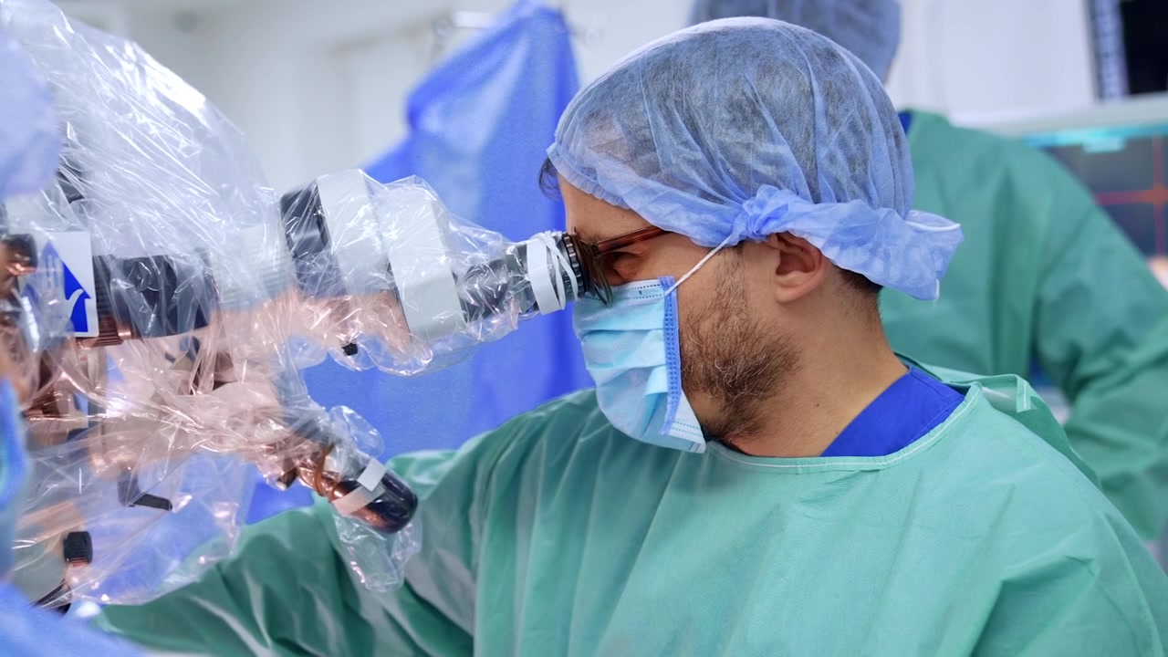 Neurocirurgião usando microscópio cirúrgico durante operação cerebral. Cirurgião com avental cirúrgico azul-petróleo, touca azul e máscara, olhando para a ocular do microscópio coberta com plástico