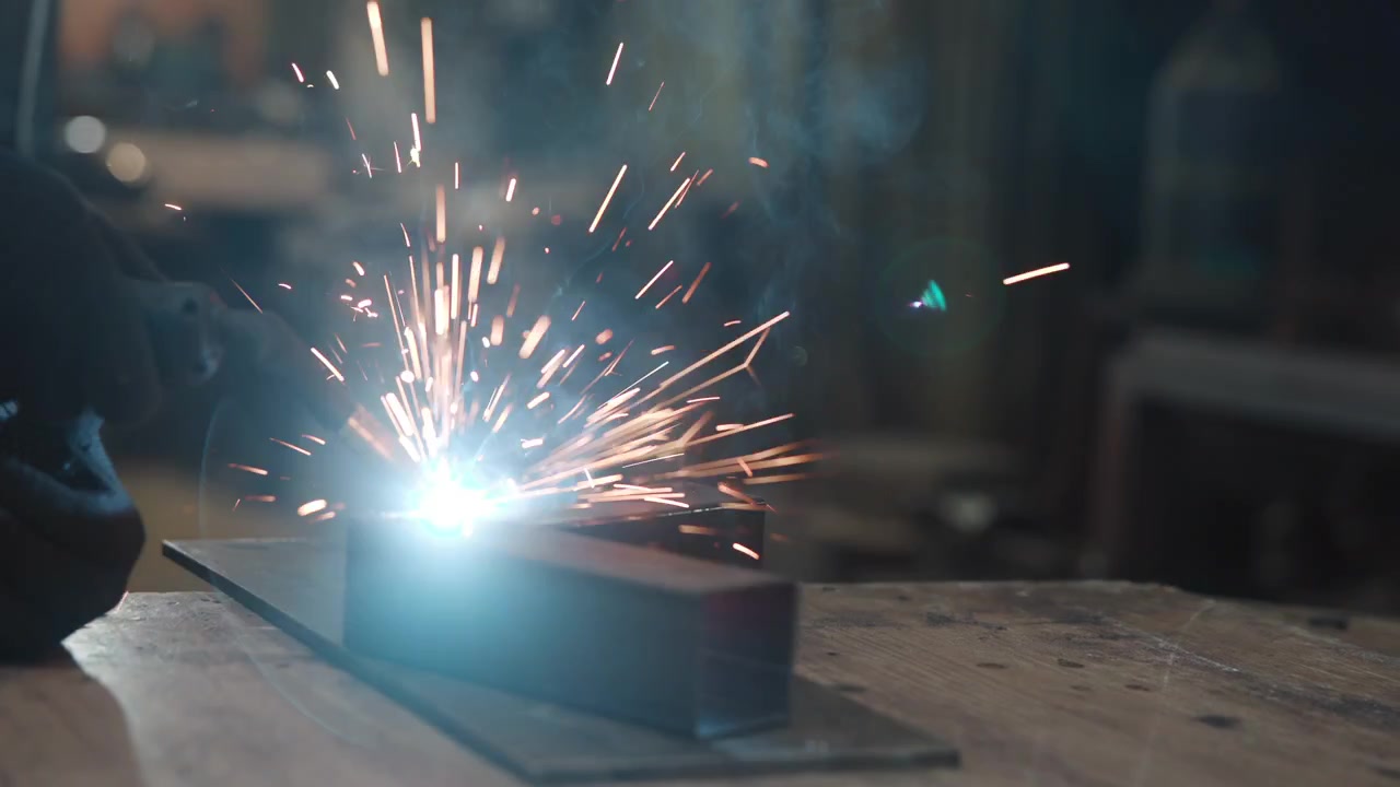 Low-angle view shows gloved hand guiding welding torch across metal joint on rectangular steel tube, with intense arc light, flying sparks, smoke, and workshop bench detail, with welding, spark, arc