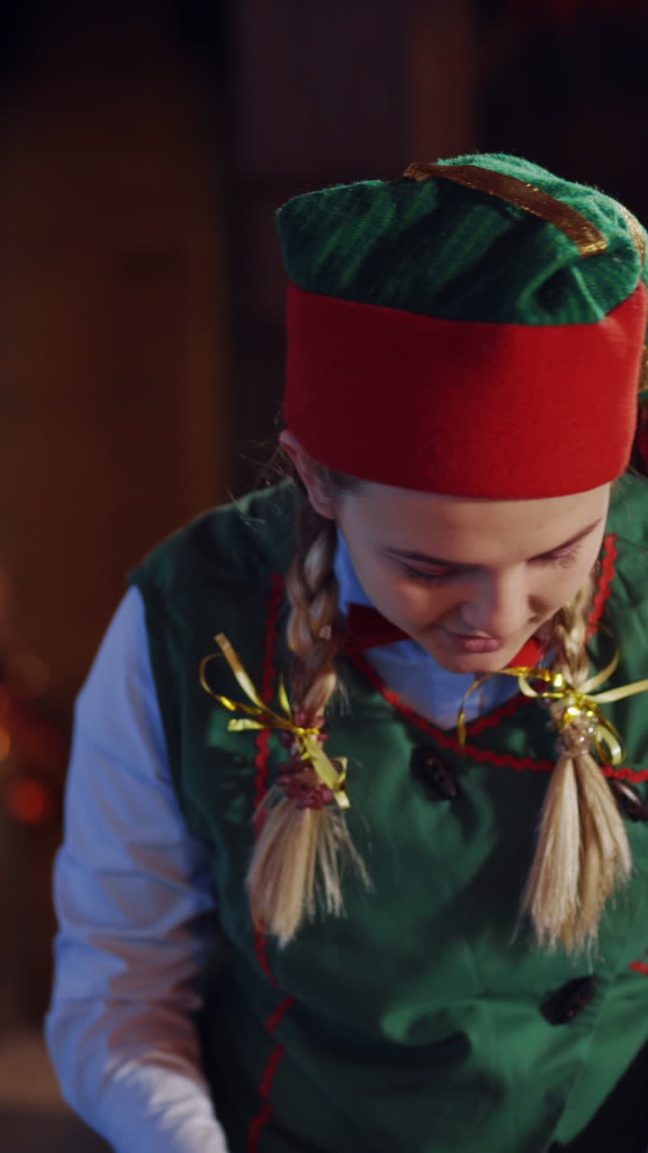 Curious elf with envelopes on the background of Christmas tree and a fireplace in the room. Happy girl in elf costume choosing envelopes and showing them to camera. Vertical video