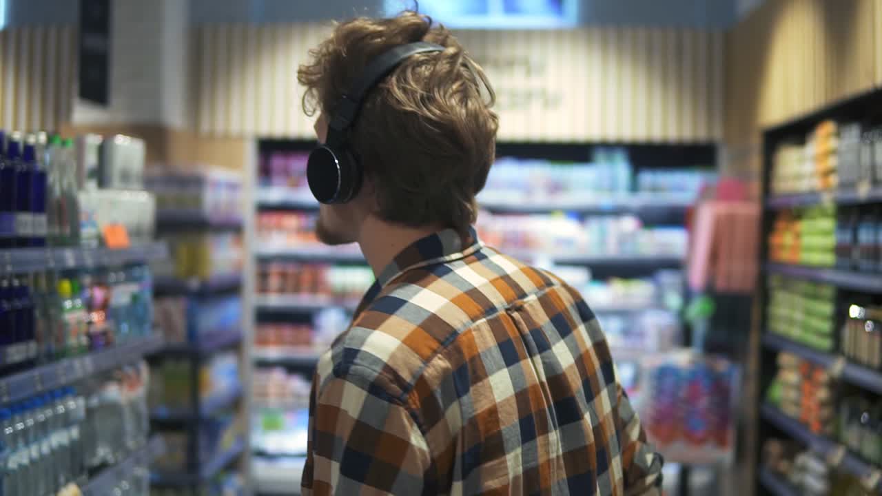 At the Supermarket: Caucasian guy with headphones walks through goods section of the store, browsing. Following back view shot