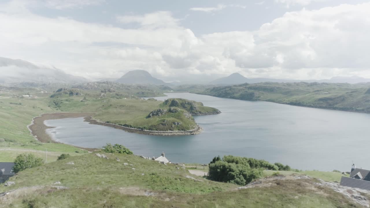 Slow drone panning shot of a Loch in Scotland with houses in the foreground, tall mountains and clouds in the distance and sun shining