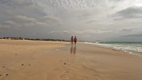 back view of romantic couple walks hand in hand along peaceful, sandy beach of boa vista island with cloudy sky overhead, creating serene and intimate moment by the ocean, cape verde