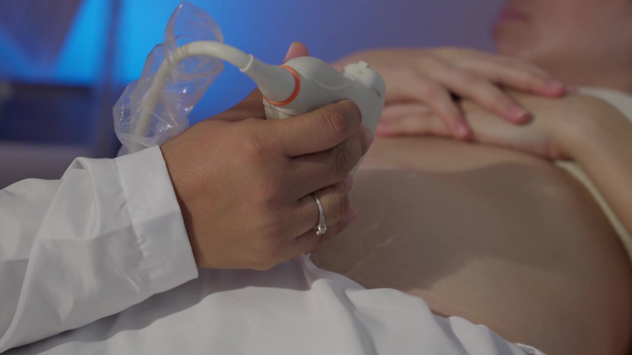 A young pregnant Latin woman lies on an examination table during a 9-month control ultrasound. The modern office features blue and purple LED lighting while a specialist operates the scanner.