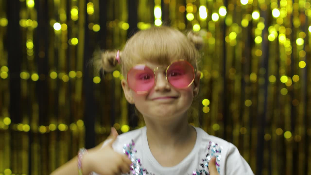 Child show thumbs up, smiling, looking at camera. Girl posing on background with foil golden curtain