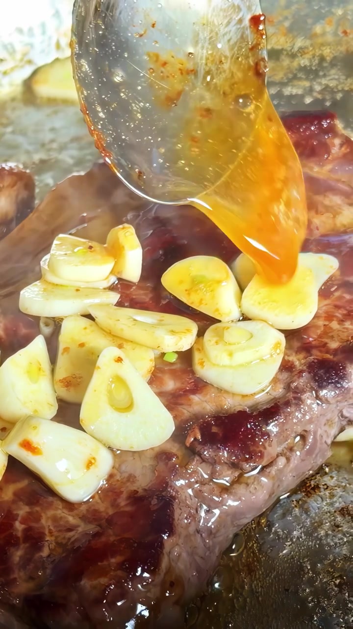 A spoon drizzles flavorful juices over a steak topped with garlic slices, enhancing its rich flavor. Perfect for cooking tutorials or culinary content.
