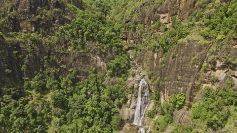 Ella-Sri-Lanka-Aerial-v43-drone-flyby-down-Ravana-Falls,-capturing-a-breathtaking-view-of-water-flowing-down-the-cliff-wall-and-the-dense-lush-jungle-vegetation---Shot-with-Mavic-3-Cine---April-2023