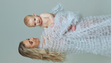 woman holding a cute lovely baby. mother wearing white robe waving a naked infant looking aside with interest. white backdrop. vertical screen.