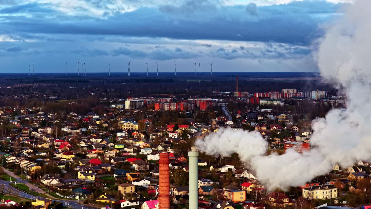 Ciminiere industriali vicino a un'area residenziale con turbine eoliche in lontananza, che illustrano il passaggio dai combustibili fossili all'energia rinnovabile all'interno di un paesaggio urbano e industriale.