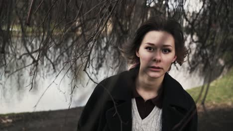 Young-woman-in-a-black-coat-standing-under-a-windy-tree-in-a-park-in-autumn-in-afternoon-3