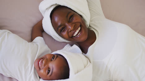 portrait of african american mother and daughter wearing towels lying on bed and smiling